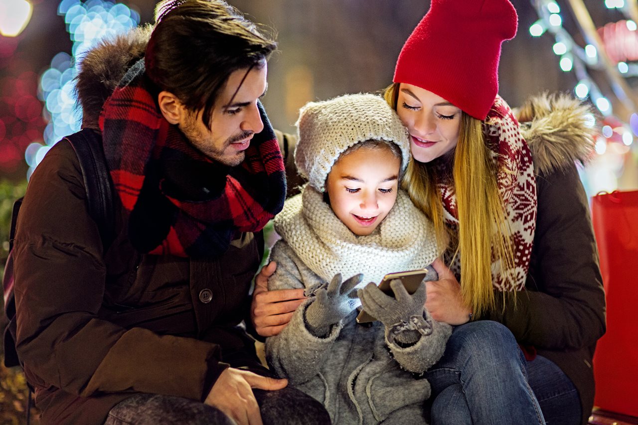 Couple and their little girl bundled up in winter wear sitting on a bench under holiday lights while video chatting with Grandma.