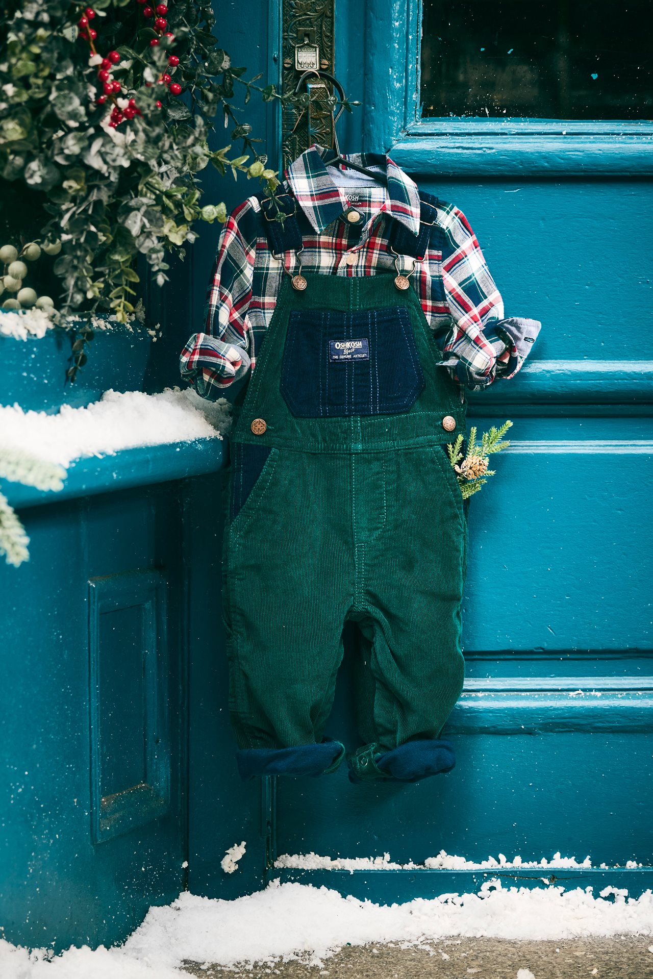 Green overalls with plaid flannel shirt on hanger next to holiday wreath with red berrys on it.