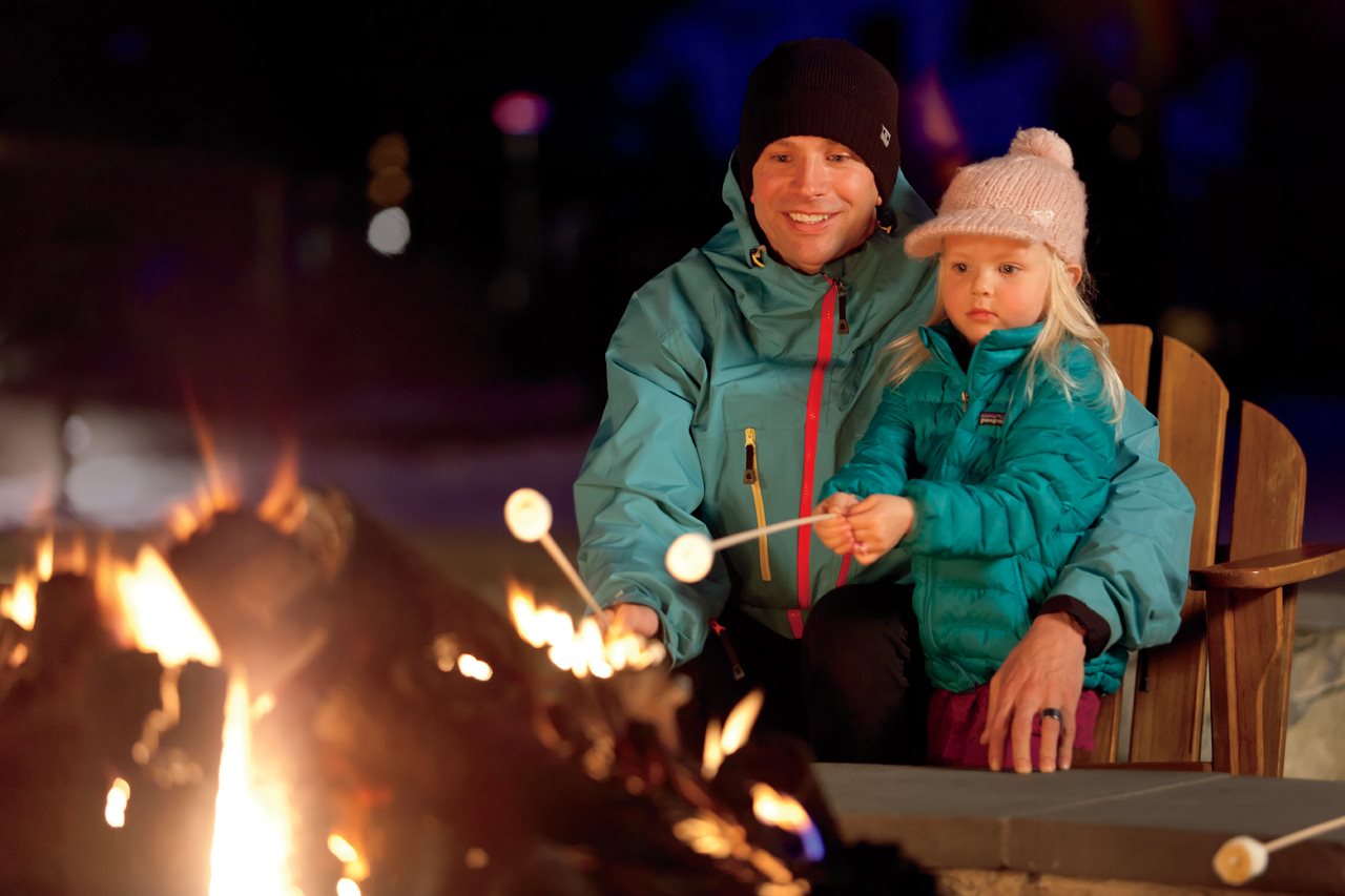 Dad and little daughter roasting marshmellows over a fire in Winter outside The Ritz Calton: Lake Tahoe.