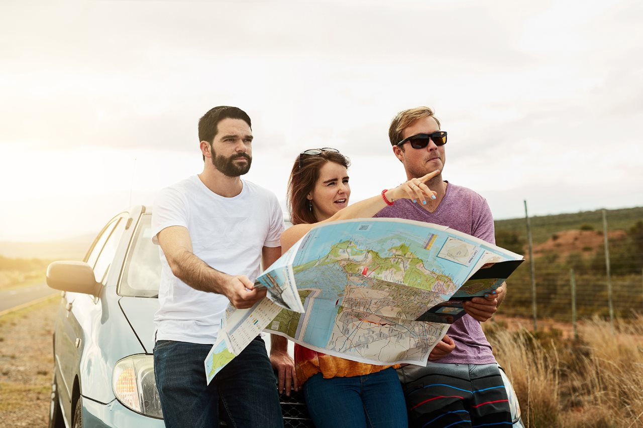 Group of three friends leaning on a car figuring out their trip using a map.