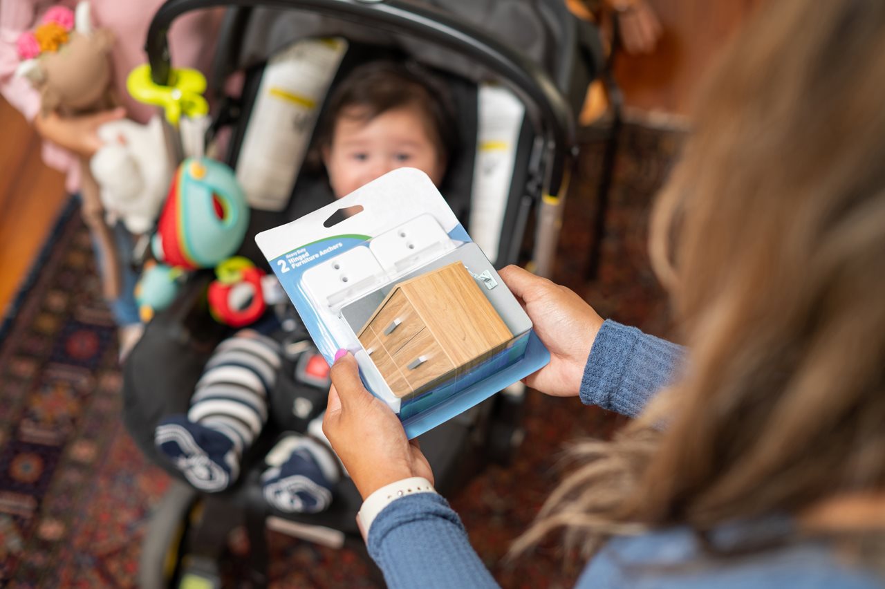 Mother holding a package to anchors used to secure a dresser to the a wall and her little baby in a car seat in the background.