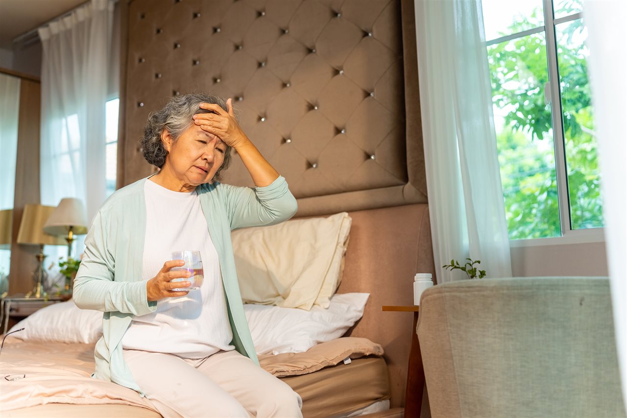 Senior woman sitting on her bed with her eyes closed and one hand on her forehead while the other hand is holding a glass of water.