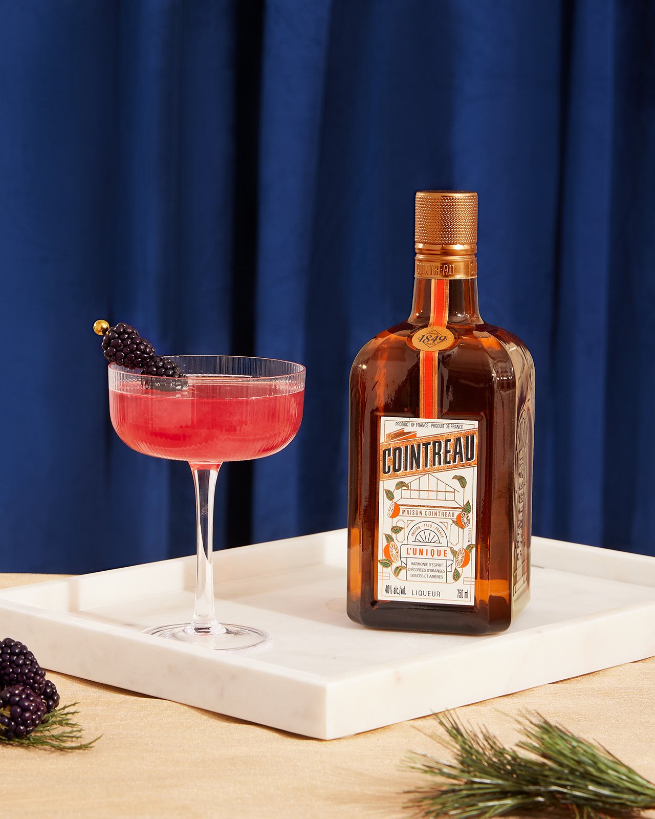 A Holiday Jam Cosmo next to a bottle of Cointreau on a white tray in front of a blue curtain.